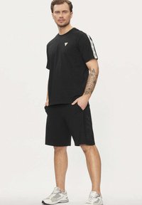 Maglietta nera in cotone con un piccolo logo triangolare, abbinata a pantaloncini neri con stampa testurizzata "GUESS" sul lato. Scarpe da ginnastica completano il look.