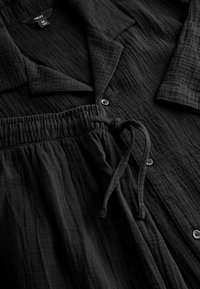 Czarny dwuczęściowy strój z koszulą zapinaną na guziki i spodniami z elastycznym pasem. Materiał ma fakturę pomarszczoną. Zawiera sznurek do regulacji.