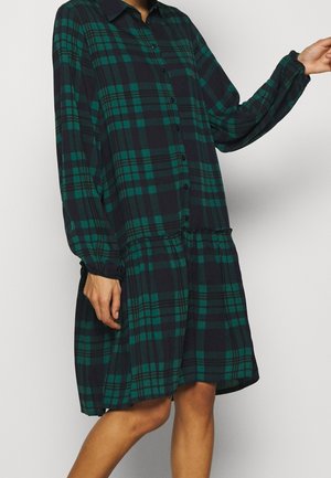 Frau trägt ein langärmliges, grün-schwarz kariertes Kleid mit Knöpfen, tiefer Taille und gerafftem Saum, steht vor einem schlichten Hintergrund.