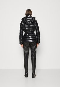 Mujer de pie, vistiendo una chaqueta acolchada brillante negra con cinturón y capucha, pantalones de cuero negros y botas de tacón negras, frente a un fondo blanco liso.