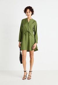 Robe verte à manches courtes avec un col en V, bouton à l'avant, taille cintrée et ourlet asymétrique. Associée avec des sandales noires à lanières et un sac à main noir.