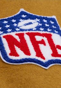 NFL-Logoaufnäher mit roten und weißen Buchstaben auf blauem Hintergrund mit weißen Sternen, auf eine braune Stoffoberfläche genäht.