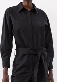 Abito camicia lungo nero con maniche lunghe in tessuto strutturato, dotato di colletto, due tasche sul petto e una cintura annodata in vita.