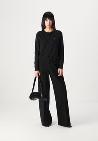 Zwarte cardigan met knopen, gedragen met wijde zwarte broek. Model houdt een zwarte gepatineerde handtas vast met een gebogen silhouet.