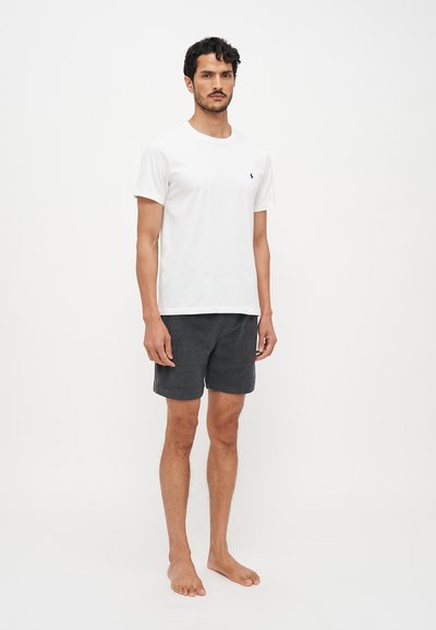 T-shirt alb din bumbac cu un logo mic pe piept, purtat cu pantaloni scurți gri închis, texturat, de lungime medie. Modelul stă desculț pe un fundal simplu.
