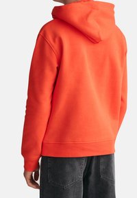 Pullover-Hoodie in leuchtendem Orange, mit einer großen Kapuze, gerippten Bündchen und einer weichen Textur. Kombiniert mit dunkelgrauen Hosen.