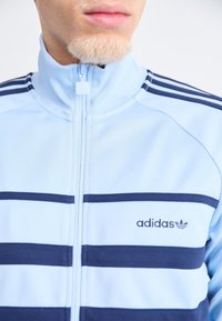Ljusblå zip-up jacka med marinblå horisontella ränder, ståkrage och broderad Adidas-logotyp på vänster bröst.