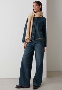 Denim jacket with a button front, paired with wide-leg jeans. A light beige scarf with fringed ends accents the outfit.