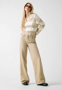 Beige Hosen mit weitem Bein, kombiniert mit einem cremefarbenen und beigen gestreiften Pullover. Das Material wirkt weich, mit einem gerippten Saum und Kragen. Sonnenbrille rundet den Look ab.