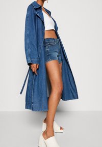 Žena v dlouhém denimovém kabátu, bílém crop topu, denimových šortkách a bílých platformových sandálech s otevřenou špičkou stojí proti jednobarevnému pozadí.