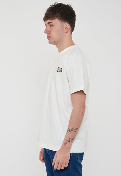 Camiseta blanca de algodón con cuello redondo, mangas cortas y un pequeño logo negro en el lado izquierdo del pecho. Llevada con jeans azules.