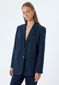Blazer in blu navy con righe sottili, colletto ampio, chiusura con un solo bottone e due tasche frontali. Realizzato in un tessuto testurizzato. Vestibilità su misura.