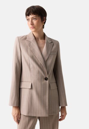 Kvinde med kort brunt hår iført beige blazer med striber og matchende bukser, der kigger til siden mod en hvid baggrund.