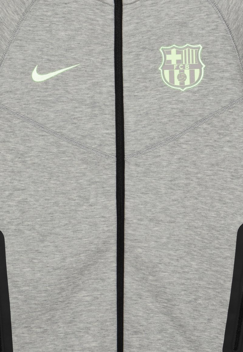 Veste zippée grise en tissu doux, avec des accents noirs et un logo FC Barcelona réfléchissant sur la poitrine. Logo Nike Swoosh en vert.