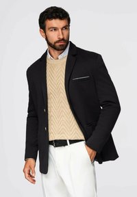 Schwarze Blazer mit taillierter Passform, beigem strukturiertem Pullover darunter, weißen Hosen und einem schwarzen Gürtel, mit einer Brusttasche als Akzent.
