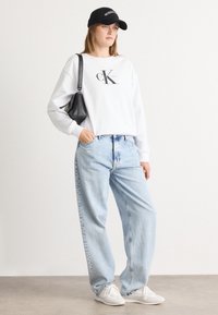 Sweat-shirt blanc avec le logo noir "Calvin Klein", jean bleu clair à coupe ample, casquette noire et baskets blanches. Sac à épaule en cuir noir.