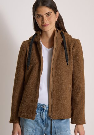 Mujer con cabello largo y oscuro que lleva una chaqueta marrón con textura y cremallera, con cordones negros, encima de una camisa blanca y jeans azules.