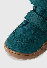Scarpa in pelle testurizzata color turchese con suola in gomma marrone e una larga fascia sulla parte superiore, mostrata in primo piano su uno sfondo bianco.