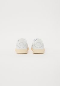 Zapatillas blancas con parte trasera redondeada, suelas de goma beige y detalles en gris claro a los lados. Textura suave con sutiles acentos de logo.