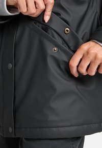 Svart vattentät jacka med en mjuk textur, som har en sidoficka med tryckknappsstängning och kontrasterande sömnadsdetaljer.