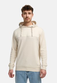 Beige hoodie med et tekstureret mønster, der har en justerbar hætte, diskret logo og ribbede manchetter og nederkant. Båret sammen med lyseblå jeans.