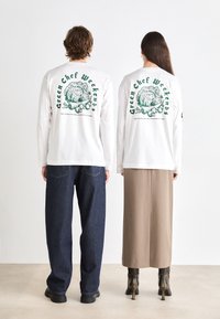Hvite langermede skjorter har grønn grafisk tekst "Green Chef Weekends" og grønnsaksdesign bak. Mørk denim- og beige skjørt bæres under.