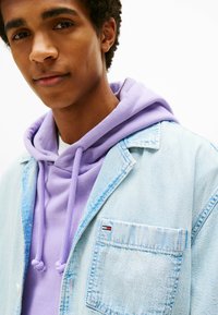 Jasnoniebieska kurtka dżinsowa nałożona na lawendowy hoodie z troczkami, z kieszenią na piersi z małym detalem logo.