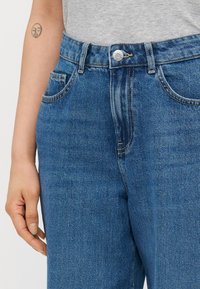 Modre jeans hlače z visokim pasom, ki imajo bakrene zakovice in šive, stranske žepke in gladko teksturo.