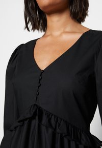 Vestido preto com decote em V, mangas bufantes, botões e uma bainha rodada. O tecido parece suave e leve, oferecendo uma silhueta estruturada.