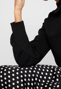 Musta pitkähihaisen puseron hihoissa on liehuke-erityiskohta, yhdistetty valkoisilla pilkuilla varustettuun mustaan polka dot -hameeseen. Sileä kangaspinta.