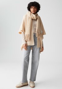 Poncho lavorato a maglia beige con orli frangiati, abbinato a una sciarpa morbida e testurizzata. Jeans dritti grigio chiaro e sneaker beige completano l'outfit.