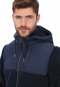 Chaqueta azul marino con cuerpo de forro polar texturizado, mangas de nylon lisas, cierre frontal con cremallera y capucha con cordón ajustable. Diseño unisex.