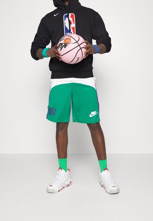 Person iført en sort NBA-hættetrøje, grønne Nike basketballshorts, grønne sokker, hvide sko, der holder en lyserød basketball med en slam dunk-grafik.