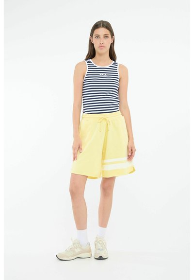 Top sin mangas a rayas en azul marino y blanco con logo, combinado con pantalones cortos amarillos con rayas blancas. El modelo lleva calcetines blancos y zapatillas de deporte beige.