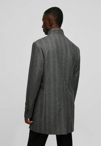 Manteau gris à motif pied-de-coq avec col haut, manches longues et coupe ajustée, présentant une texture subtile et des accents de boutons sur les manches.