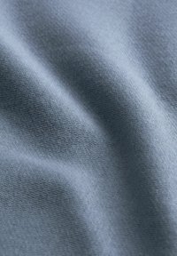 Vaaleansininen kangas, jossa on pehmeä kudottu pinta ja lempeät taitteet, jotka luovat hienovaraisia varjoja ja korostuksia.