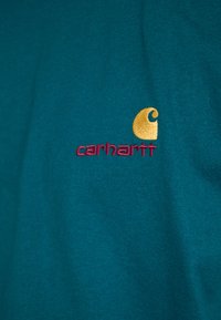 Teal cotton t-shirt featuring a yellow and maroon embroidered logo. Simple design with a short sleeve and crew neckline.