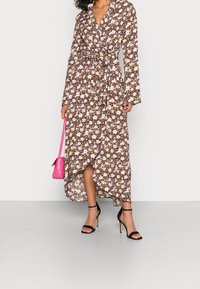 Vestido envolvente floral en marrón con flores blancas, naranjas y moradas; mangas largas, cintura con lazo, dobladillo asimétrico, combinado con tacones negros y un bolso rosa.