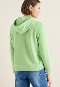 Lichtgroene hoodie met een ontspannen pasvorm, capuchon met trekkoord en geribbelde boorden. Gecombineerd met blauwe jeans, wat een casual stijl benadrukt.