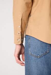Lys brun langermet skjorte med hvite knapper ved siden av en hånd og blå denim bukselomme, vist fra bak.
