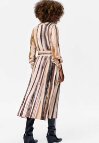 Lange, gestreepte jurk met variërende tinten beige, bruin en goud, met een aansluitende taille, lange mouwen en knopenaccenten. Draagt met zwarte laarzen.