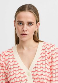 Cardigan en tricot crème avec un motif ondulé rouge, décolleté en V et fermeture à boutons. Présente un tissu texturé et des manches bouffantes.
