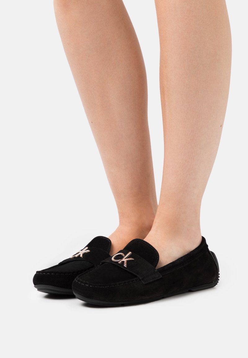 Mocassins en cuir suédé noir avec une semelle plate, présentant une lanière décorative et un logo surélevé à l'avant. Le design est simple et épuré.