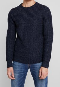 Laivastonsininen neulepaita, jossa on teksturoitu ribbineulos, pyöreä kaula ja pitkät hihat, yhdistetty sinisiin farkkuihin. Paksu kangas ja vartalonmyötäinen siluetti.