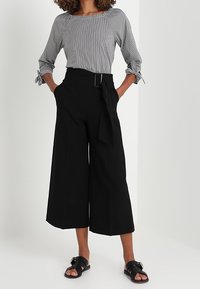 Pantaloni neri a gamba larga con vita regolabile, abbinati a una blusa a righe grigie e bianche. I sandali hanno un dettaglio con fibbia.