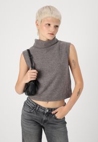 Mladý človek s krátkymi blond vlasmi, oblečený v bezrukávovom šedom roláku, má čiernu kabelku na ramene a šedé džínsy, pričom jednu ruku má v vrecku.