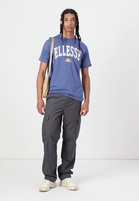 Mėlynas trumpomis rankovėmis marškinėliai su balta "Ellesse" logotipu, derinami su pilkais kargo džinsais su keliais kišenėmis ir bežė sportinėmis bateliais.