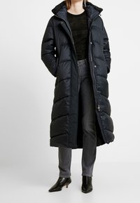 Long black puffer coat with a hood and textured inner lining. Features a zip closure and button accents, paired with dark jeans and heeled boots.