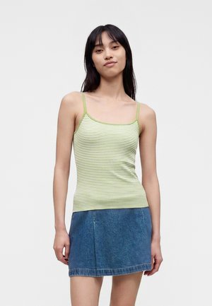 Ung kvinde iført en lysegrøn og hvidstribet tanktop med tynde stropper og en blå denim nederdel, stående mod en simpel baggrund.