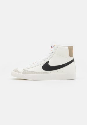 Biały wysoki sneakers Nike z czarnym logo, beżowymi akcentami na pięcie i palcach oraz białymi sznurowadłami na jednolitym jasnoszarym tle.